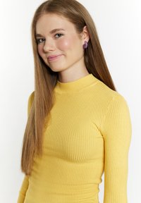 Gelbes, geripptes Turtleneck-Oberteil aus dehnbarem Material, kombiniert mit runden lila Ohrringen. Langes, glattes Haar, das hinabfällt.