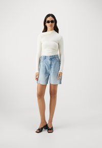 Lettvekts, langermet kremtop kombinert med falmet blå denimshorts. Tilbehør inkluderer svarte solbriller og glansede hælsandaler.
