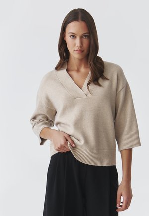 Beige, gebreide sweater met een V-hals, geribde mouwen en rand. Losse pasvorm en zachte textuur. Gekoord over zwarte broeken.