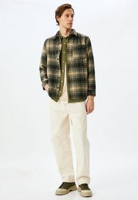 Chemise en flanelle à carreaux dans des tons verts et beiges, superposée sur une chemise olive, combinée avec un pantalon crème et des chaussures kaki.