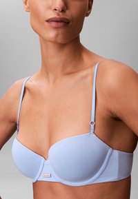 Sujetador acolchado azul claro con tirantes ajustables, tejido liso, soporte con aro y una etiqueta con logo en la banda. Diseño sencillo y moderno.