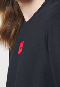 Maglione nero in cotone con scollatura rotonda. Presenta una toppa quadrata rossa con il testo "HUGO". Texture liscia e design aderente.