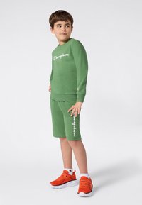 Garçon portant un sweat à capuche Champion vert et un short avec des baskets orange, debout en regardant à droite sur un fond blanc uni.