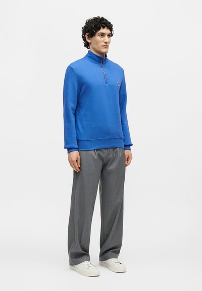 Άνδρας που στέκεται, φοράει ένα φωτεινό μπλε πουλόβερ με φερμουάρ στο τέταρτο, γκρι φαρδιά παντελόνια και λευκά αθλητικά παπούτσια σε λευκό φόντο.