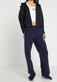 Chaqueta con capucha negra con bolsillos frontales combinada con pantalones de chándal azul marino que presentan franjas laterales blancas y un parche de logo. Calzado negro.