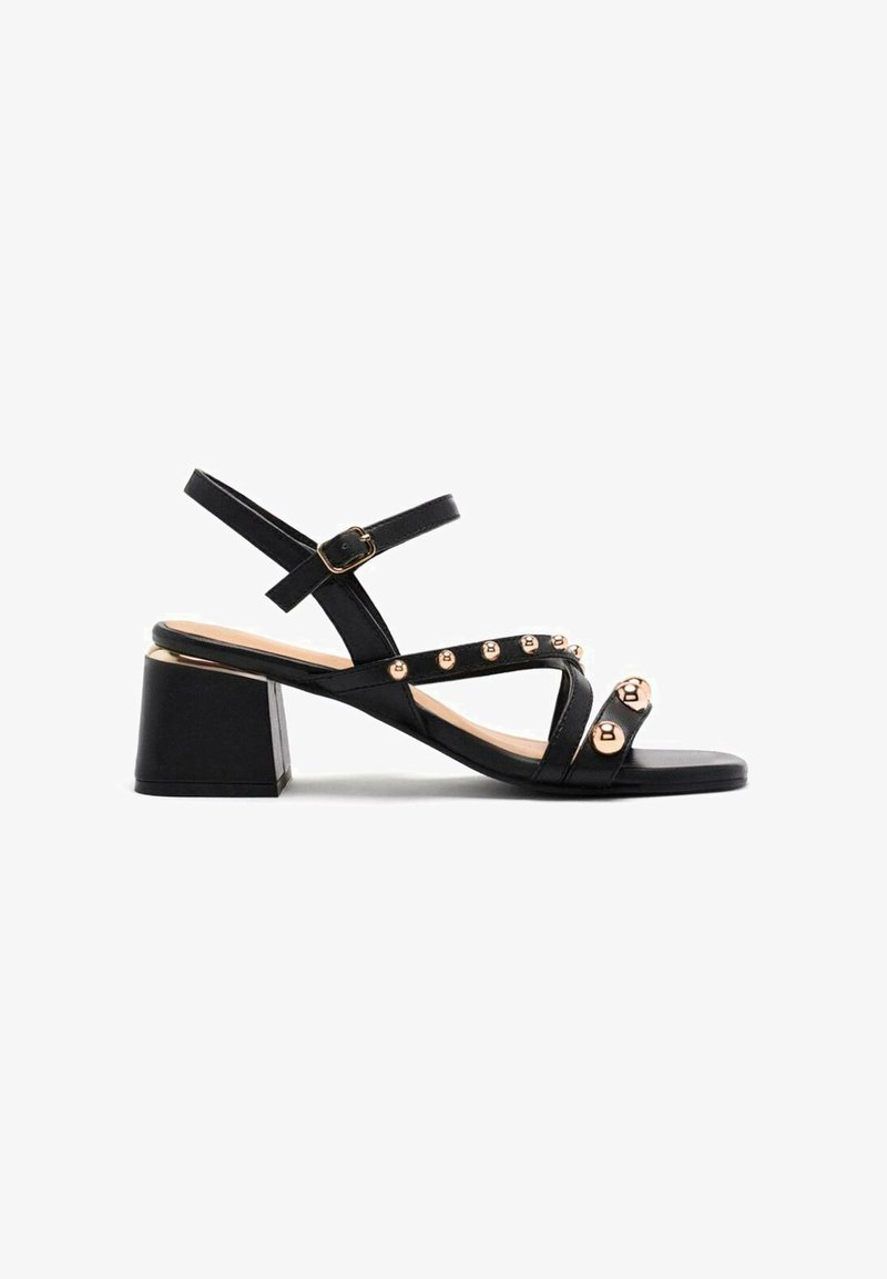 Sandalias de tacón negro con tacón bloque, que cuentan con múltiples correas con detalles en tono dorado y tachuelas redondas, y una textura de cuero suave.