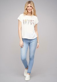 T-shirt blanc à manches courtes avec imprimé floral, associé à un jean skinny bleu clair et des baskets blanches. Design simple et décontracté.