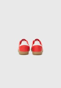 Röda syntetiska sneakers med vita hälaccenter och sulor av gummigummi, med en slät textur och rundad design.