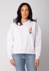 Grijze katoenen sweatshirt met drop shoulders, met daarop een kleurrijke geborduurde gnoom op de linkerborst. Gecombineerd met blauwe spijkerbroek.