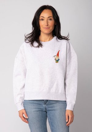 Grijze katoenen sweatshirt met drop shoulders, met daarop een kleurrijke geborduurde gnoom op de linkerborst. Gecombineerd met blauwe spijkerbroek.