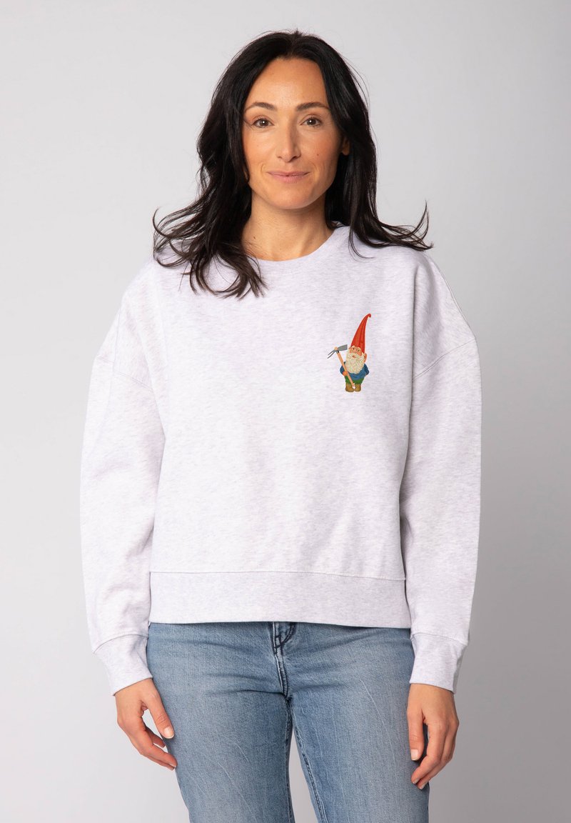 Grijze katoenen sweatshirt met drop shoulders, met daarop een kleurrijke geborduurde gnoom op de linkerborst. Gecombineerd met blauwe spijkerbroek.