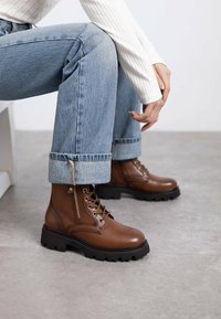 Botas de cuero marrón hasta el tobillo con una suela negra gruesa, cordones en la parte frontal y cremallera lateral. Combinadas con jeans de mezclilla azul claros y un top blanco de canalé.