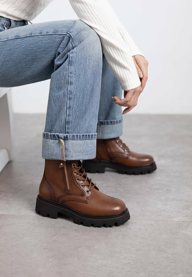 Botas de cuero marrón hasta el tobillo con una suela negra gruesa, cordones en la parte frontal y cremallera lateral. Combinadas con jeans de mezclilla azul claros y un top blanco de canalé.