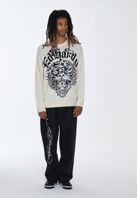 Maglione lavorato a maglia crema con grafica di un tigre nera e testo; abbinato a jeans a gamba larga neri con branding in corsivo bianco.