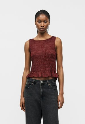 Žena s opletenými vlasmi, oblečená v bezrukávovom bordovom textúrovanom topu s volánikovým okrajom a vysokým pásom vyblednutých čiernych džínsov na sivom pozadí.
