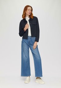 Marineblauwe cropped jas met twee voorzakken en knoopsluiting, gecombineerd met lichtblauwe wijde jeans en witte sneakers op een effen achtergrond.