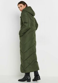 Długa oliwkowozielona kurtka puchowa z kapturem, z ukośnym pikowaniem, dwoma bocznymi kieszeniami i zamek błyskawiczny. Całość dopełniają czarne botki za kostkę.