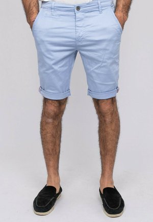 Homme portant un short bleu clair retroussé et des chaussures noires sans lacets, debout les mains dans les poches devant un fond uni.