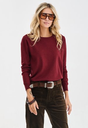 Bordeaux gekleurde langmouwtrui met een ronde hals, gecombineerd met donkerbruine corduroy broek. Accessoires bestaan uit een bruine riem en dikke armbanden.