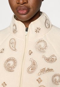 Φλις μπουφάν σε κρέμ χρώμα με ψηλό γιακά, που διαθέτει περίπλοκα καφετιά μπορντούρα και λουλουδάτη κεντητική, καθώς και μπροστινή φερμουάρ.