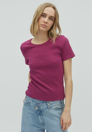 Tânără cu păr blond până la umeri, purtând un top magenta mulat cu mâneci scurte și o fustă din denim bleu asimetrică.