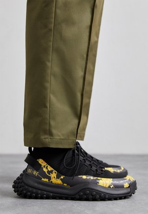 Zapatillas con patrón en negro y amarillo, suela texturizada, combinadas con pantalones verde oliva. Presentan un diseño curvo y cierre de cordones.