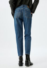 Jeans de mezclilla azul oscuro, de corte ajustado, de cintura alta, con dos bolsillos traseros, tela texturizada, con una etiqueta de marca de cuero marrón sobre la cinturilla.