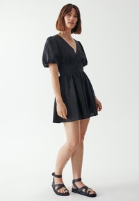 Schwarzes Kleid mit tiefem V-Ausschnitt, Puffärmeln und tailliertem Schnitt, das sich zu einem Rock ausbreitet. Genossen mit schwarzen Riemchensandalen vor einem hellen Hintergrund.