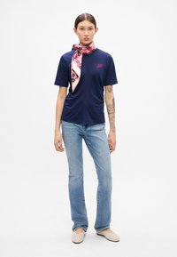 T-shirt blu scuro con maniche corte e una sciarpa a fantasia floreale legata al collo. Jeans svasati blu chiaro e scarpe slip-on beige.