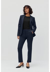 Marineblazer und taillierte Hose mit glatter Textur, kombiniert mit einem schwarzen Oberteil. Die Schuhe sind elegante schwarze Absätze mit spitzem Zeh.