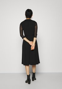 Frau mit kurzem Haar, die ein schwarzes Midikleid mit durchsichtigen Ärmeln und schwarzen Ankle Boots trägt, steht vor einer schlichten, hellgrauen Wand.