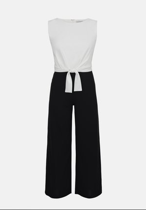 Haut blanc sans manches à nouer sur le devant, associé à un pantalon noir taille haute à jambes larges, sur un fond uni.