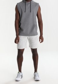 Hombre vestido con una sudadera sin mangas gris, pantalones cortos gris claro y zapatillas deportivas blancas, de pie contra un fondo blanco.