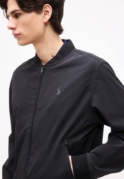 Νεαρό άτομο με σκούρα μαλλιά, φοράει μαύρο μπουφάν με φερμουάρ και μικρό κεντητό λογότυπο στο στήθος, με το χέρι στην τσέπη.