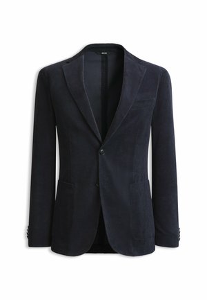 Homme portant un blazer bleu marine, une chemise à carreaux bleue, un pantalon beige et des chaussures noires, debout les mains dans les poches sur un fond blanc.