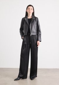 Una donna si presenta con sicurezza in una giacca di pelle cropped e pantaloni palazzo neri in satin, con uno sfondo neutro che mette in risalto il suo outfit.