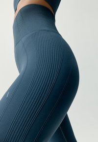 Zbliżenie osoby w wysokich, fakturowanych niebieskich legginsach bezszwowych na jednolitym jasnym tle.