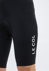 Detailný záber na čierne cyklistické šortky s vypchávkou na sedacej časti a bielym logom "LE COL" na pravej stehne na svetlom pozadí.