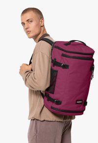 Batoh v tmavě burgundské látce s černými akcenty, se nastavitelnými popruhy, boční síťovanou kapsou a vrchní rukojetí pro snadné nošení.