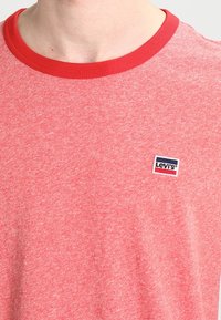 Camiseta de manga corta color rojo heather con cuello redondo y cuello rojo. Presenta un pequeño parche de logo de Levi's en azul y blanco en el pecho.