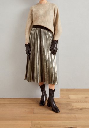 Femme portant un pull beige, une jupe midi plissée métallique, des gants en cuir marron foncé et des bottines à talons noires, debout sur un sol en bois.