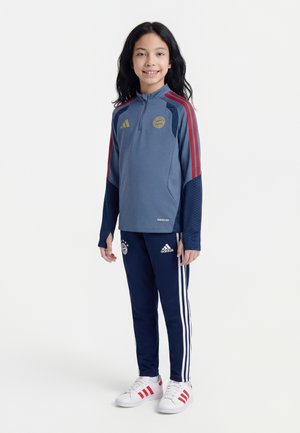 Lachend kind met lang donker haar, gekleed in een blauw-rood Bayern München sportjack, donkerblauwe adidas trainingsbroek en witte sneakers.