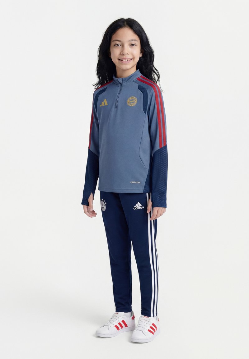 Hymyilevä lapsi, jolla on pitkät tummat hiukset, pukeutuneena sinipunaisiin Bayern Münchenin urheilutakkiin, tummansinisiin adidas-housuihin ja valkoisiin lenkkitossuihin.