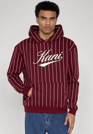 Ung mand iført en vinrød hættetrøje med hvide lodrette striber og teksten "Karri", stående med den ene hånd i forlommen og blå jeans.