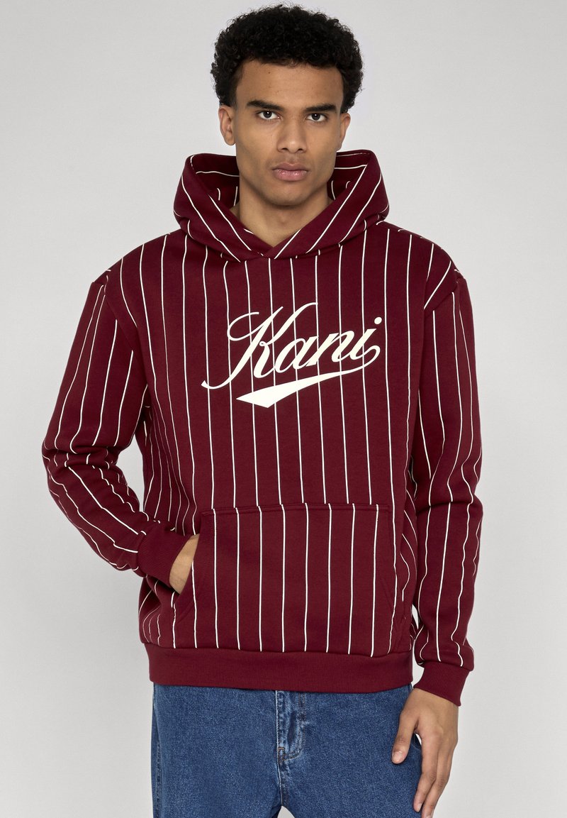 Jeune homme portant un sweat à capuche bordeaux avec des rayures verticales blanches et le texte "Karri", debout avec une main dans la poche avant et un jean bleu.