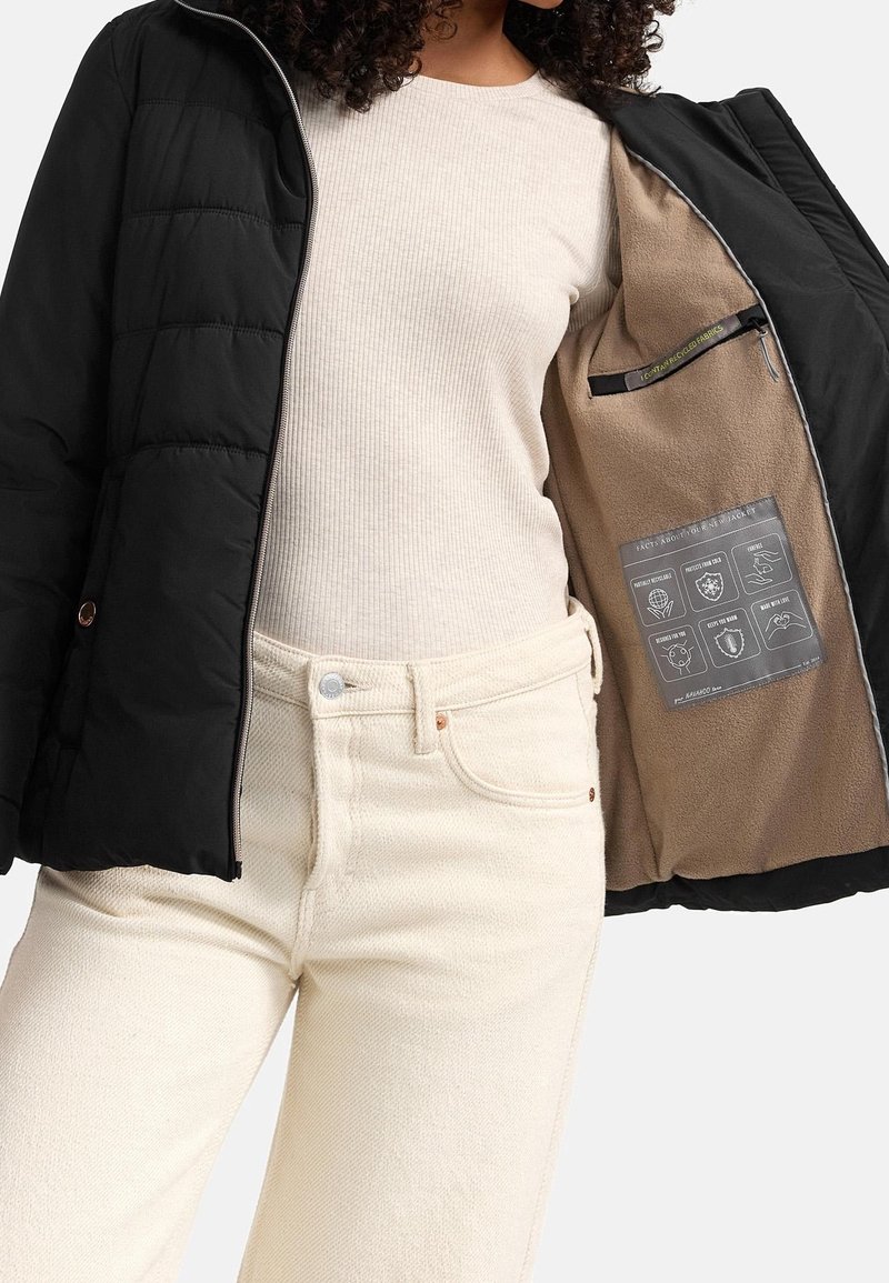 Černá péřová bunda s útulným béžovým interiérem, se zapínáním na zip a etiketou s pokyny uvnitř. Model má na sobě světlé džíny a top.