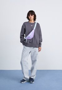 Paarse en lichtblauwe schoudertas gedragen over een grijze sweatshirt, gecombineerd met lichte spijkerbroek en donkere sneakers, tegen een eenvoudige achtergrond.