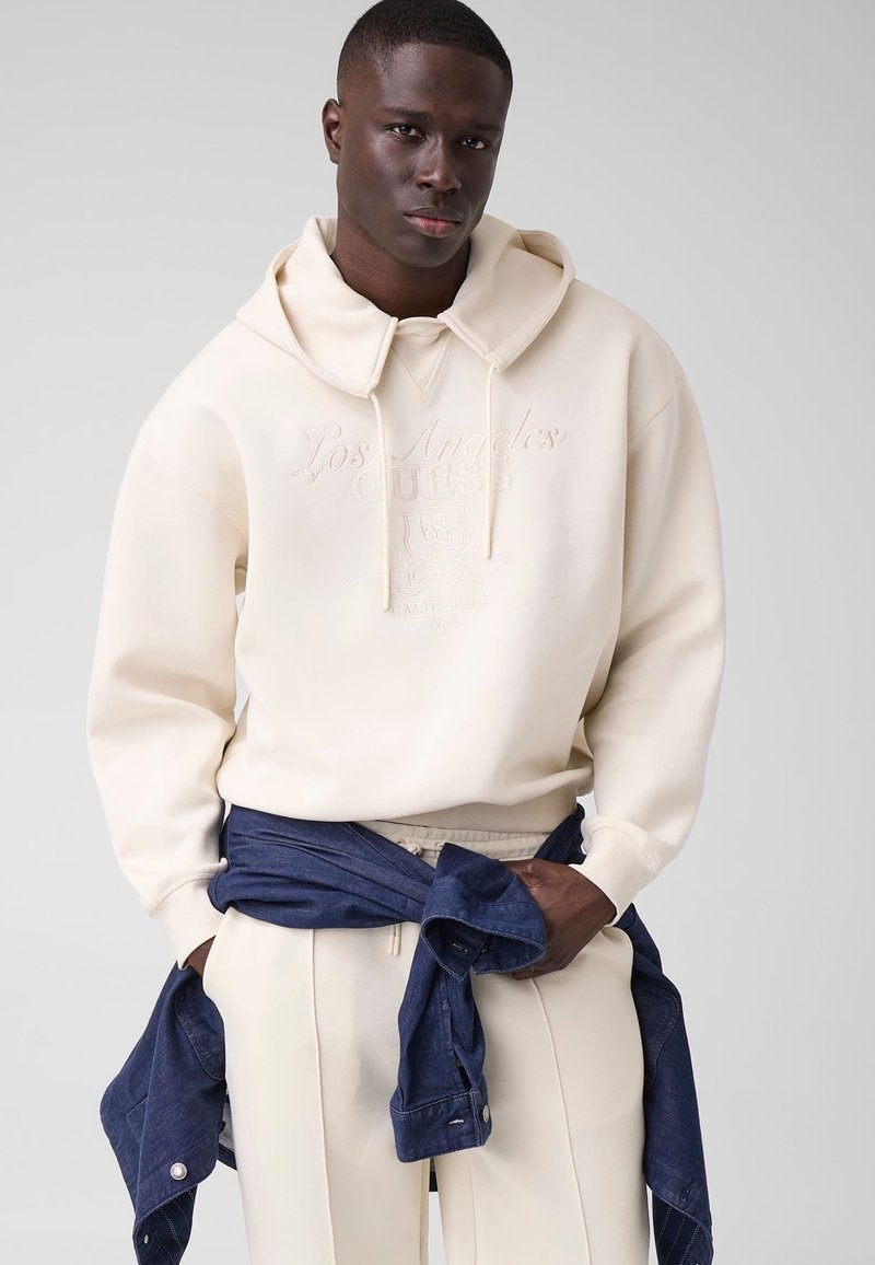 Homme portant un sweat à capuche crème avec le texte "Los Angeles", un pantalon crème et une veste bleu foncé nouée autour de sa taille sur un fond uni.