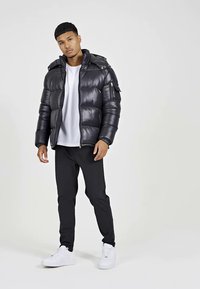 Zwarte gewatteerde jas met een capuchon, ritssluiting en zijzakken. Gecombineerd met zwarte broek en witte sneakers, tegen een eentonige achtergrond.