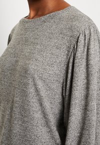 Primo piano di una persona che indossa un maglione a maglia grigia, con una scollatura rotonda e dettagli arricciati sulle spalle.